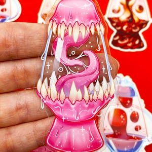 Pu&ograve; includere: Un adesivo rosa trasparente con un design stilizzato di lampada a lava con bocca e lingua. L'adesivo ha denti bianchi e un effetto gocciolante, con altri adesivi di lampada a lava sullo sfondo.