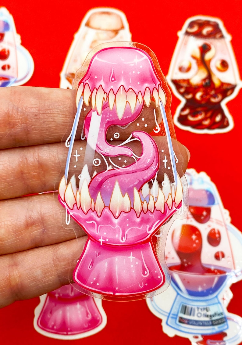 Pu&ograve; includere: Un adesivo rosa trasparente con un design stilizzato di lampada a lava con bocca e lingua. L'adesivo ha denti bianchi e un effetto gocciolante, con altri adesivi di lampada a lava sullo sfondo.