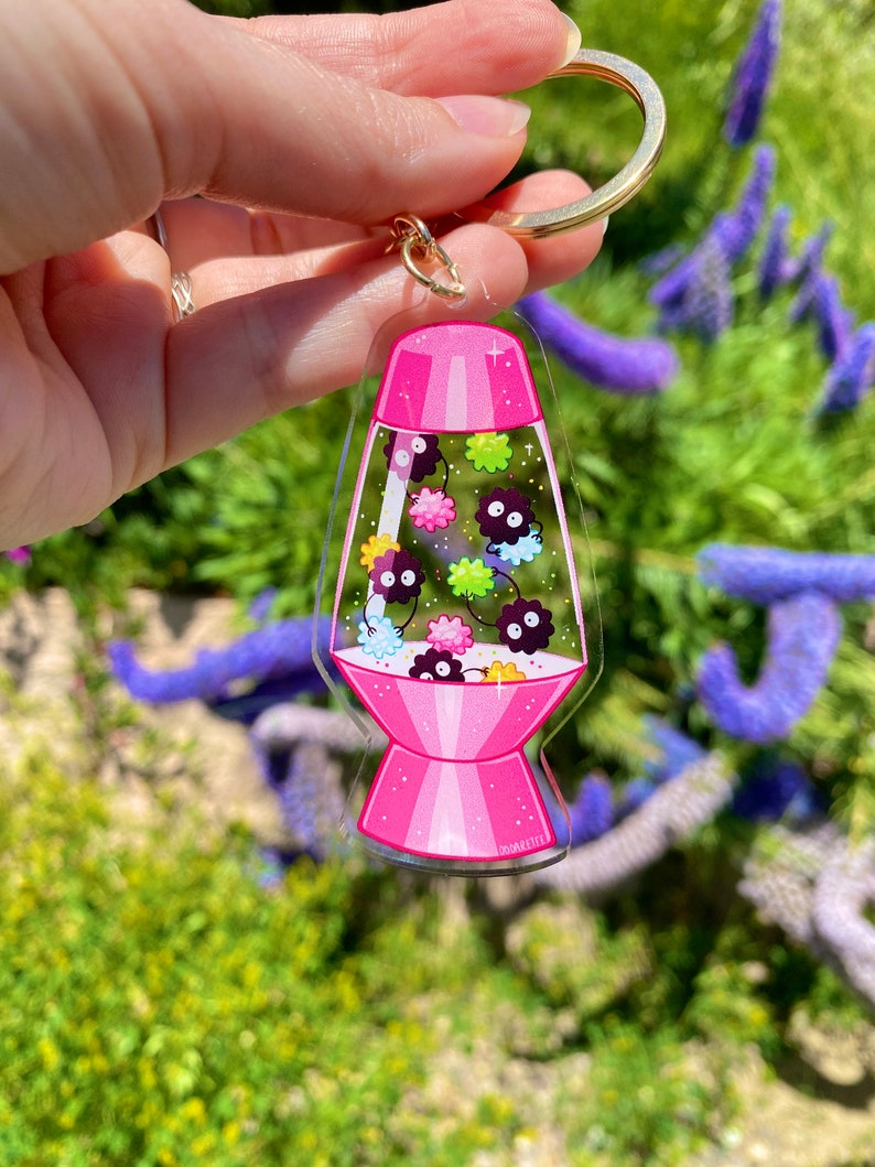 May include: A pink and clear acrylic keychain shaped like a lava lamp with a gold key ring. The lamp contains small black and white creatures with pink, green, and yellow accents.