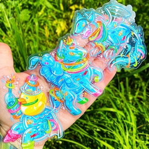May include: Three clear stickers featuring blue cartoon creatures with rainbow rings and glitter. The creatures are a unicorn, a dragon, and a duck. The stickers are on a hand against a green background.