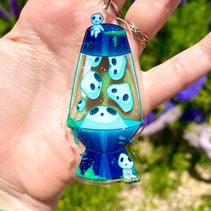 May include: A clear acrylic keychain shaped like a lava lamp with a blue and green design. The lamp is filled with white ghosts and has a small white ghost sitting on top. The keychain has the text "Oddaretie" on the bottom.