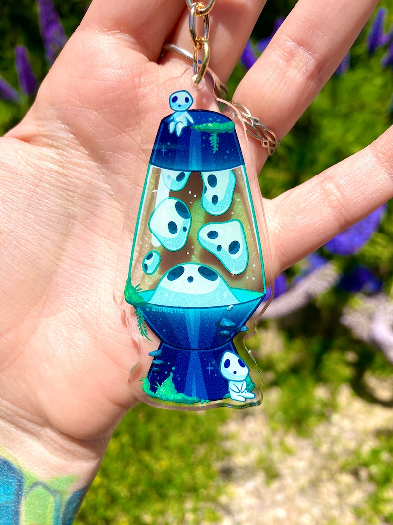May include: A clear acrylic keychain shaped like a lava lamp with a blue and green design. The lamp is filled with white ghosts and has a small white ghost sitting on top. The keychain has the text "Oddaretie" on the bottom.