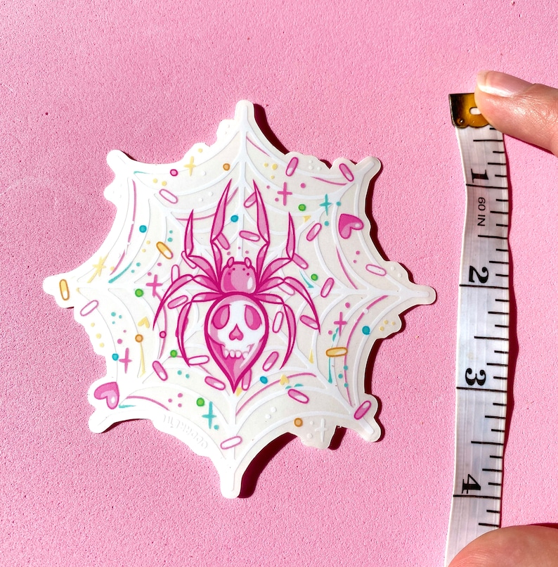 May include: Pink spider with a skull face in a white web with colorful confetti and hearts. The sticker is approximately 3 inches in diameter.