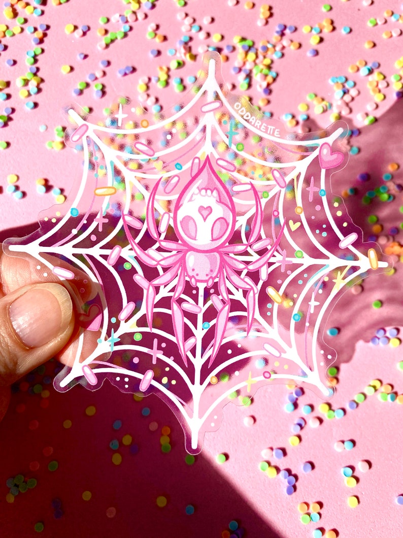 May include: A pink spider with a heart on its chest is trapped in a white web with pink, blue, and yellow sprinkles. The text "ODDARETTE" is visible in the upper right corner of the image.