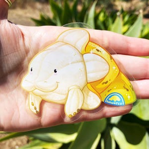 K&ouml;nnte beinhalten: Ein transparenter Aufkleber mit einem Cartoon-Fisch in Wei&szlig; und Gelb mit glitzernder Textur. Der Fisch hat einen kleinen Mund und gro&szlig;e, runde Augen. Der Aufkleber tr&auml;gt den Text "Oddarete" und "Inawatee".