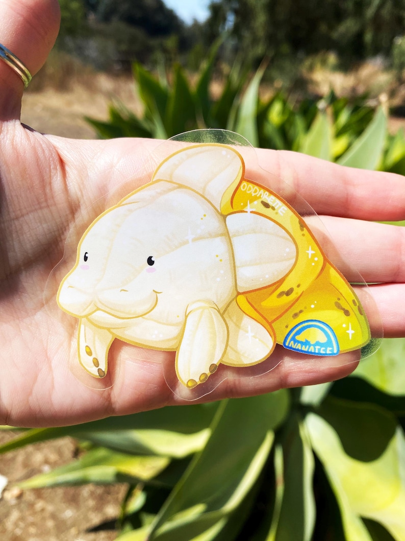 K&ouml;nnte beinhalten: Ein transparenter Aufkleber mit einem Cartoon-Fisch in Wei&szlig; und Gelb mit glitzernder Textur. Der Fisch hat einen kleinen Mund und gro&szlig;e, runde Augen. Der Aufkleber tr&auml;gt den Text "Oddarete" und "Inawatee".