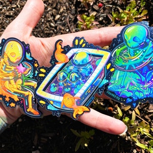May include: Three iridescent holographic stickers featuring skeletal figures in various poses. The stickers are on a hand with a tattoo. The stickers are on a dark background with stars and other details. The text "ODDARETTE" is visible on one of the stickers.