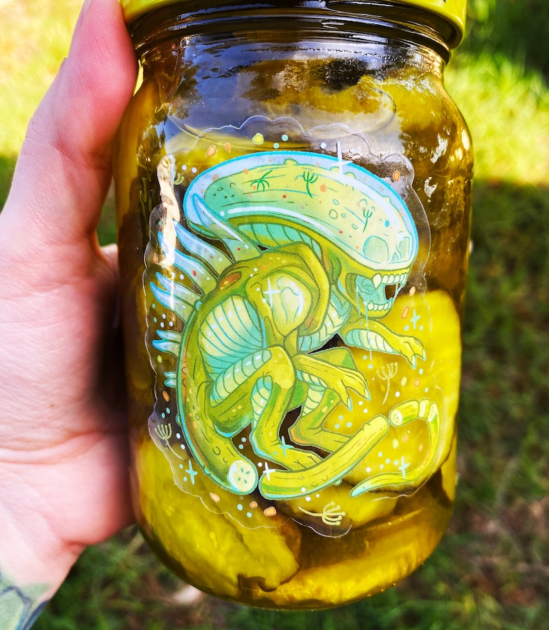 K&ouml;nnte beinhalten: Ein Glas gef&uuml;llt mit gelben Gurken, mit einem Aufkleber eines gr&uuml;nen Au&szlig;erirdischen mit einem gro&szlig;en Kopf und einem langen Schwanz.