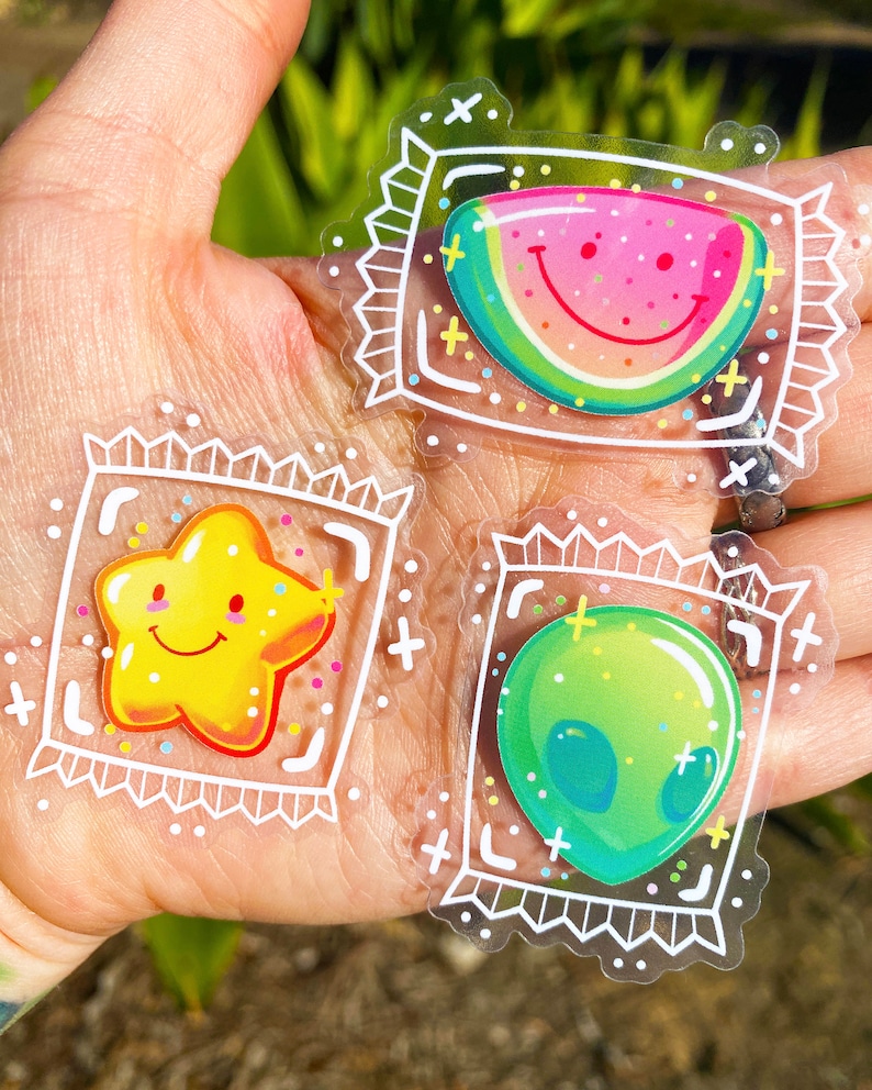 Puede incluir: Tres pegatinas transparentes con contornos blancos e ilustraciones coloridas en su interior. Las pegatinas representan una sand&iacute;a sonriente, una estrella sonriente y un extraterrestre verde. Cada pegatina tiene un borde blanco con un patr&oacute;n repetitivo de signos m&aacute;s y &aacute;ngulos rectos.