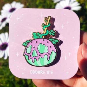 May include: A green and pink enamel pin shaped like a watermelon with a skull face and a green stem. The pin is on a pink card with white stars and the text "Oddaretite".