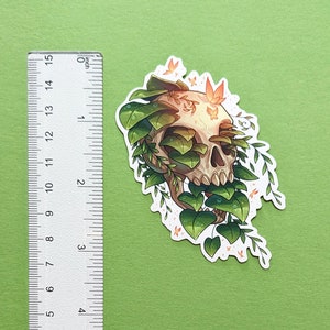 May include: A sticker featuring a white skull with green vines and leaves growing over it. The skull has a detailed design with visible teeth and eye sockets. There are small orange butterflies scattered around the skull.