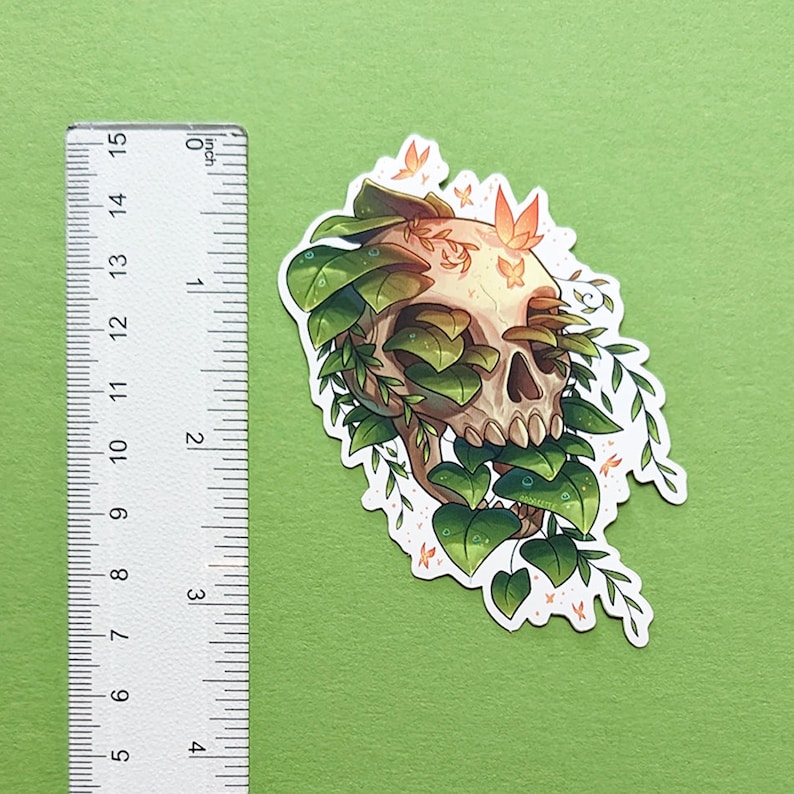 May include: A sticker featuring a white skull with green vines and leaves growing over it. The skull has a detailed design with visible teeth and eye sockets. There are small orange butterflies scattered around the skull.