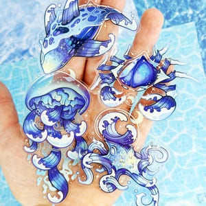 May include: A set of four clear stickers featuring stylized marine creatures including a whale, a jellyfish, a crab, and an octopus. Each creature is rendered in shades of blue and white with wave-like patterns, sparkling accents, and small crescent moon details.