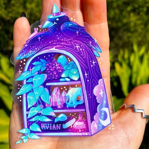 May include: A clear sticker featuring a purple raven wearing a witch hat, perched atop a shelf filled with crystals, a skull, and plants. The word "AVIAN" is printed on the shelf. The sticker has a whimsical, magical theme.
