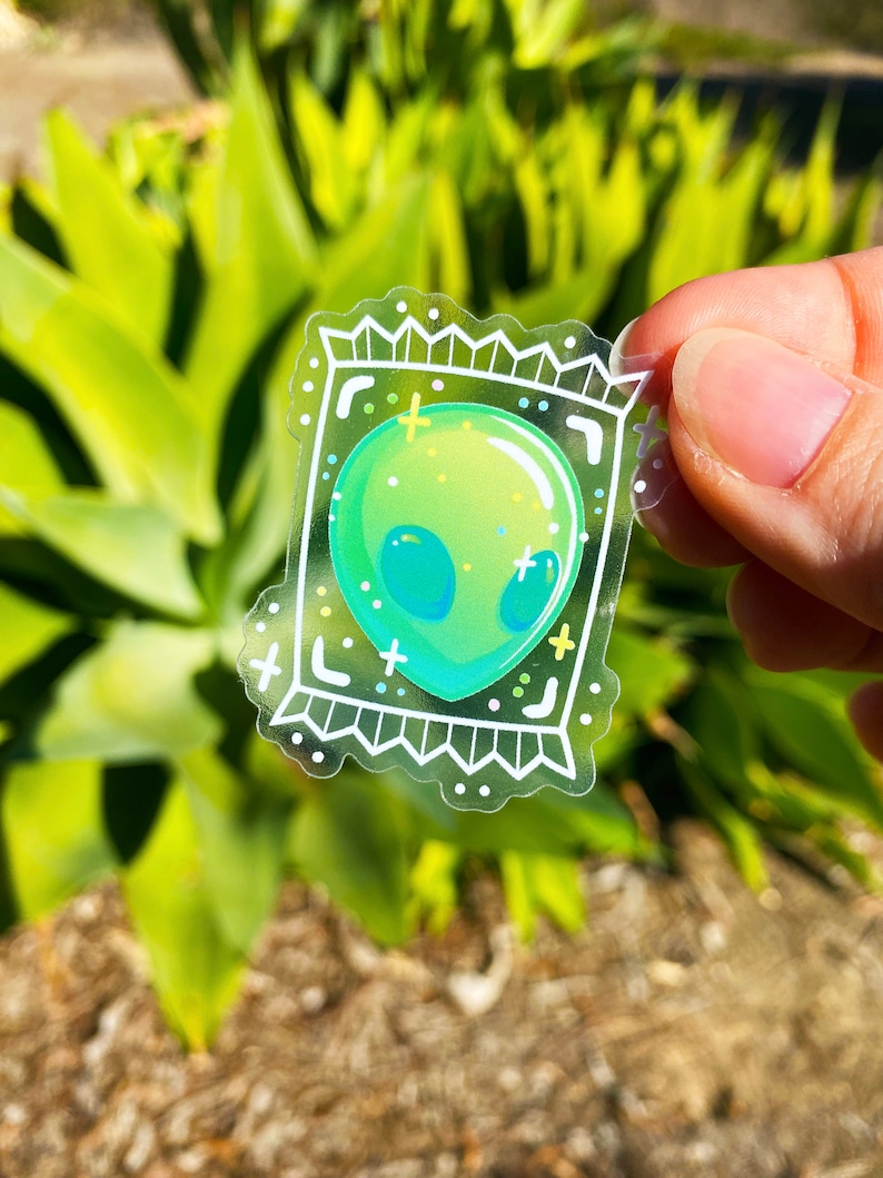 Puede incluir: Una pegatina transparente con una cabeza de extraterrestre verde dentro de un marco blanco. El extraterrestre tiene un cuerpo verde con ojos azules y un contorno blanco. El marco tiene un borde blanco con un patr&oacute;n blanco y negro.