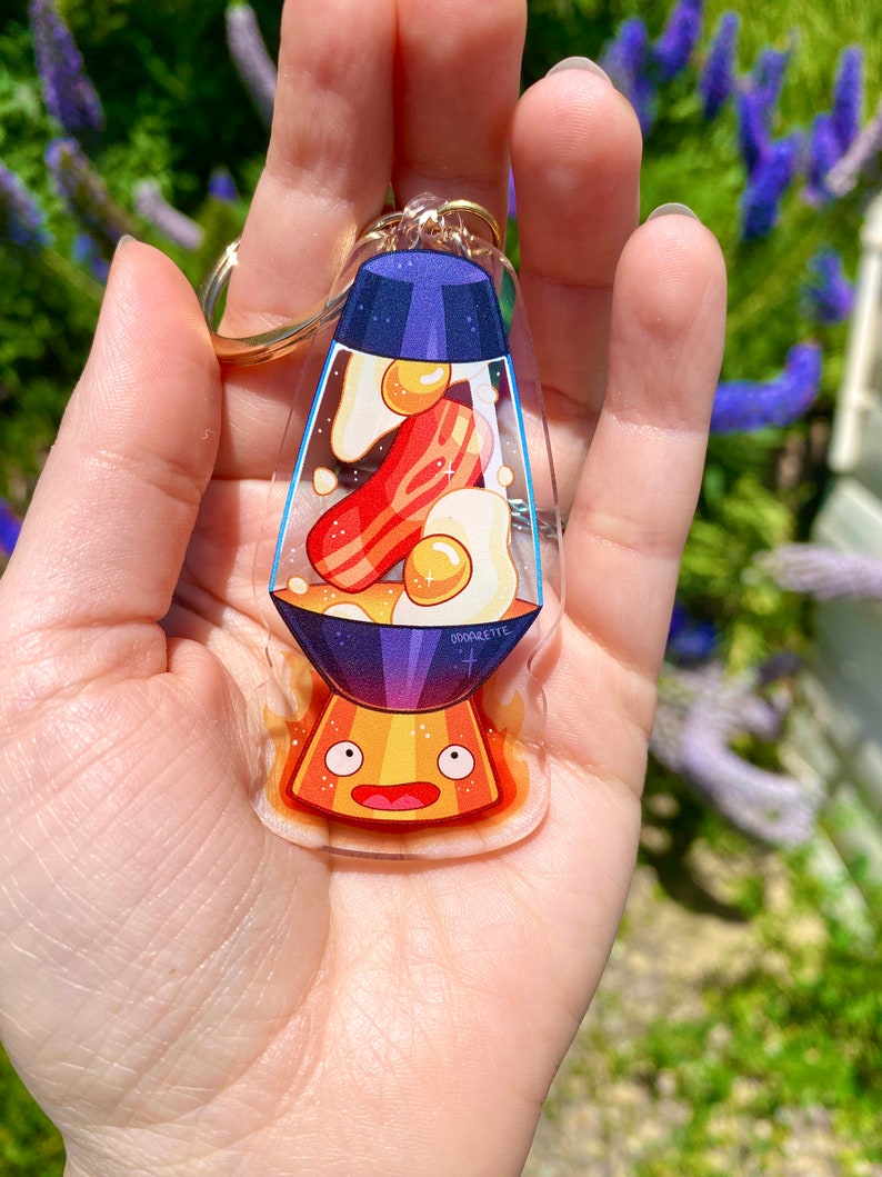 May include: A clear glass keychain shaped like a lava lamp with a blue top and a yellow base. Inside the lamp are cartoon images of bacon and eggs. The base of the lamp has a cartoon face with eyes and a mouth. The text "GOODNIGHTTE" is printed on the lamp.