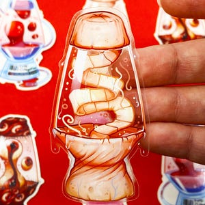 Pu&ograve; includere: Un adesivo trasparente con un design a forma di lampada a lava a fumetti con un interno a forma di gamberetti e gelatina. L'adesivo ha una combinazione di colori rosso, arancione e rosa. Altri adesivi con design simili sono visibili sullo sfondo.