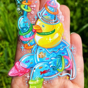 May include: A yellow rubber ducky wearing a blue and green hat with a ring toss game around it. The ducky is holding a blue and green ring toss stick. The ducky is surrounded by blue water with fish and other rings. The sticker has a sparkly finish and says "Oddareite" at the bottom.
