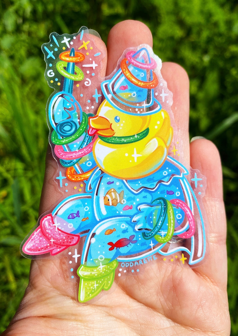 May include: A yellow rubber ducky wearing a blue and green hat with a ring toss game around it. The ducky is holding a blue and green ring toss stick. The ducky is surrounded by blue water with fish and other rings. The sticker has a sparkly finish and says "Oddareite" at the bottom.