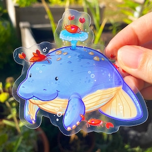 May include: A clear sticker featuring a blue manatee with a yellow tail fin and three red crabs. The manatee is swimming in a blue and white ocean with bubbles. The sticker has the text "ODDPARADISE" in the bottom right corner.