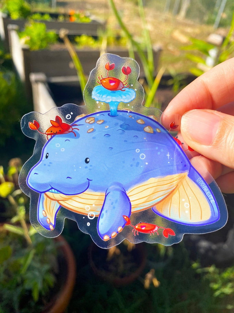 May include: A clear sticker featuring a blue manatee with a yellow tail fin and three red crabs. The manatee is swimming in a blue and white ocean with bubbles. The sticker has the text "ODDPARADISE" in the bottom right corner.