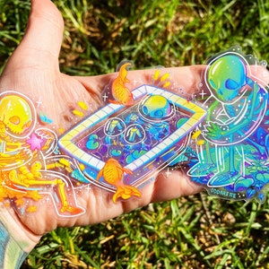 May include: Three clear holographic stickers featuring skeletons in various poses. One skeleton is holding a star, another is sitting in a pool with fish, and the third is standing in a garden. The stickers are on a hand with green grass in the background. The text "ODDARETE" is visible on the sticker.