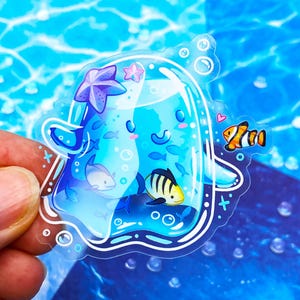 May include: A clear sticker featuring a cartoon jellyfish with a smiling face, surrounded by small fish, a starfish, and bubbles. The jellyfish is a light blue color, and the sticker has a white outline.