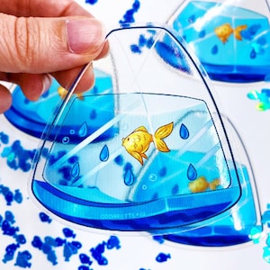 May include: A clear plastic sticker with a blue and white illustration of a goldfish in a triangular fishbowl. The sticker is surrounded by blue fish-shaped glitter.
