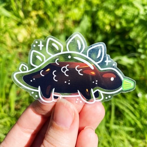 K&ouml;nnte beinhalten: Ein transparenter Aufkleber mit einer schwarzen und wei&szlig;en Umrandung eines Cartoon-Stegosaurus. Der Stegosaurus hat ein gr&uuml;nes Blatt am Schwanz und eine wei&szlig;e Umrandung eines gepunkteten Musters um ihn herum.