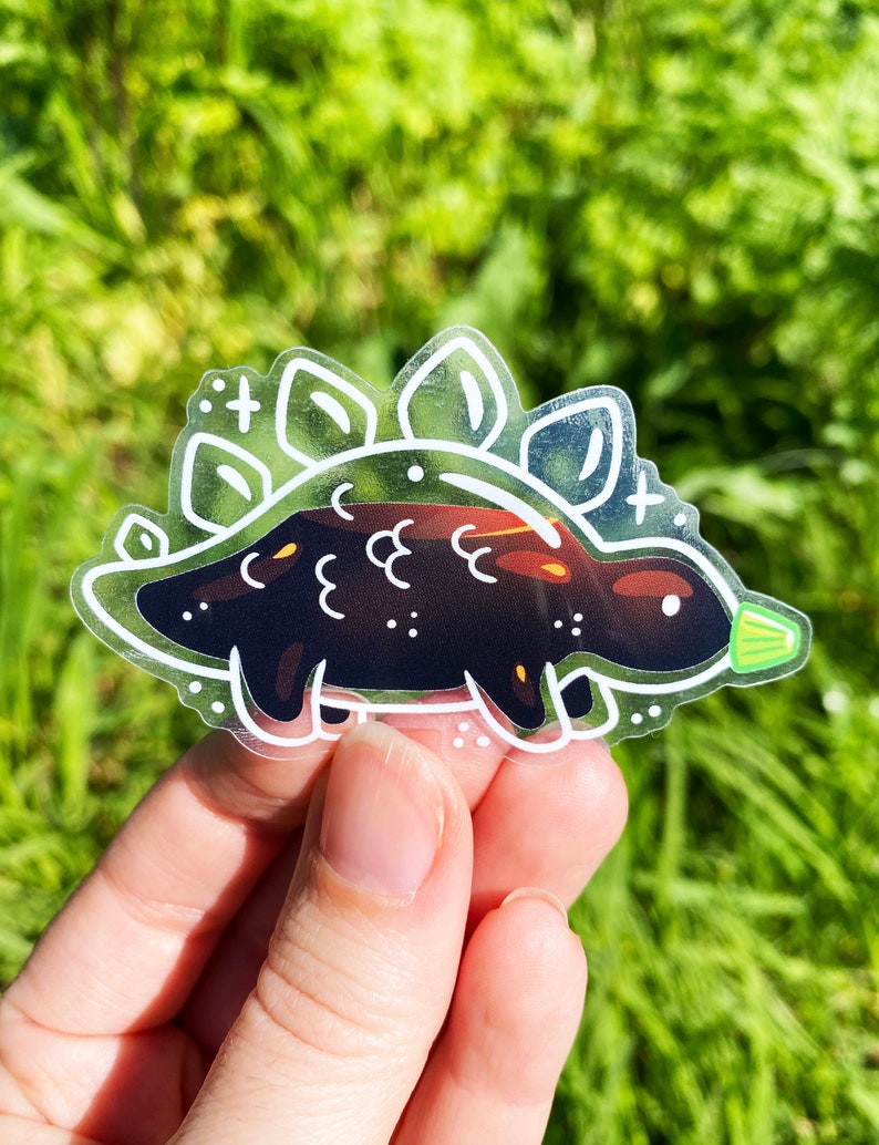 K&ouml;nnte beinhalten: Ein transparenter Aufkleber mit einer schwarzen und wei&szlig;en Umrandung eines Cartoon-Stegosaurus. Der Stegosaurus hat ein gr&uuml;nes Blatt am Schwanz und eine wei&szlig;e Umrandung eines gepunkteten Musters um ihn herum.