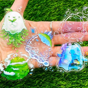 May include: A collection of clear, whimsical stickers featuring various aquatic and nature-themed designs. The stickers include a white jellyfish with green accents, a blue and green island, and a blue creature with a starfish on its head.