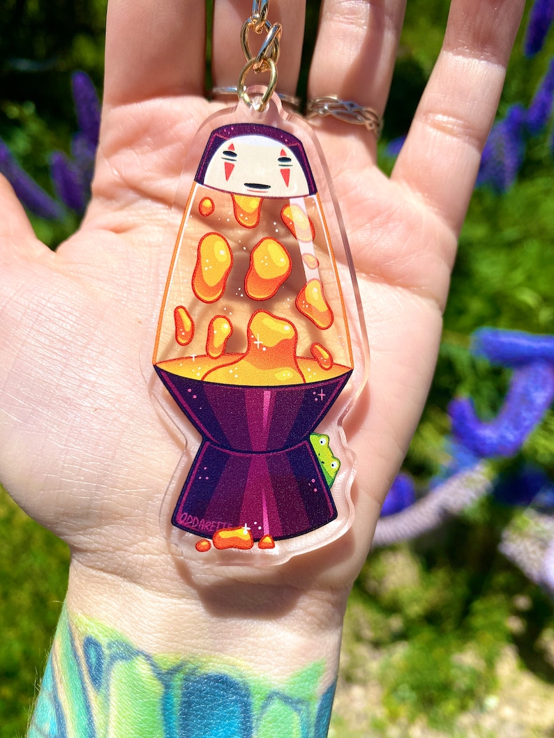 May include: A clear acrylic keychain shaped like a lava lamp with a cartoon face inside. The lava is orange and yellow and the base is purple. The keychain has the text "OddAret" on the base.