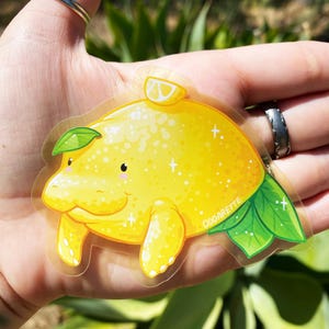 K&ouml;nnte beinhalten: Ein transparenter Aufkleber mit einem Cartoon-Manati in Form einer Zitrone. Der Manati ist gelb mit einem gr&uuml;nen Blatt auf dem Kopf und einer Zitronenscheibe oben drauf. Der Aufkleber hat eine glitzernde Textur und die Worte "OddArette" sind unten rechts aufgedruckt.