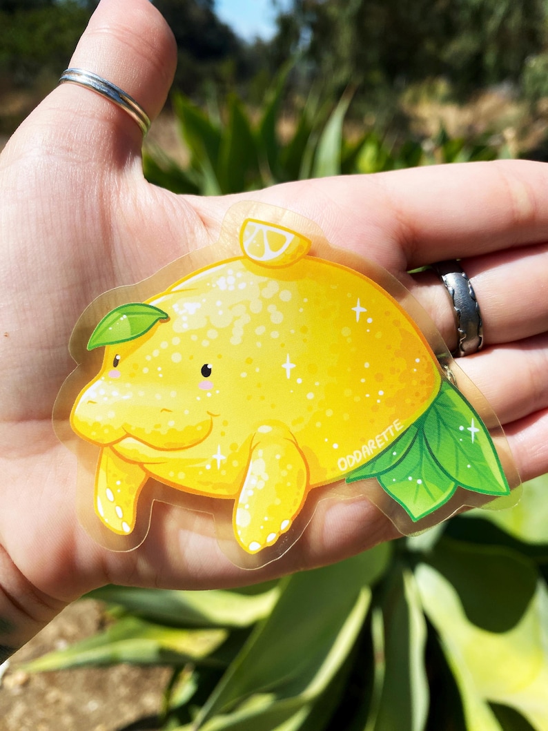 K&ouml;nnte beinhalten: Ein transparenter Aufkleber mit einem Cartoon-Manati in Form einer Zitrone. Der Manati ist gelb mit einem gr&uuml;nen Blatt auf dem Kopf und einer Zitronenscheibe oben drauf. Der Aufkleber hat eine glitzernde Textur und die Worte "OddArette" sind unten rechts aufgedruckt.