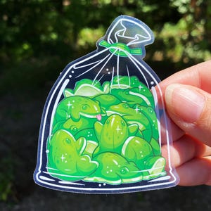 May include: A clear sticker featuring a bag filled with cartoon green frogs. The frogs have white highlights and some have their tongues sticking out. The bag is tied with a green ribbon and has a white outline.