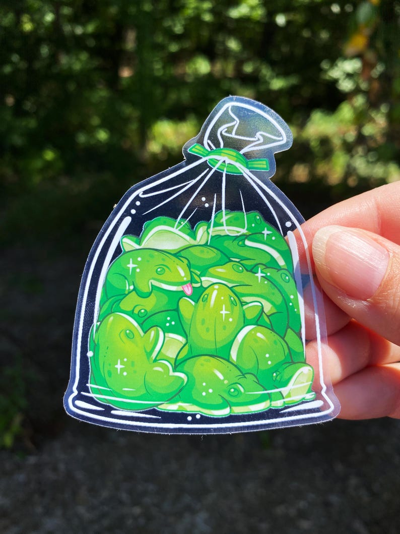 May include: A clear sticker featuring a bag filled with cartoon green frogs. The frogs have white highlights and some have their tongues sticking out. The bag is tied with a green ribbon and has a white outline.
