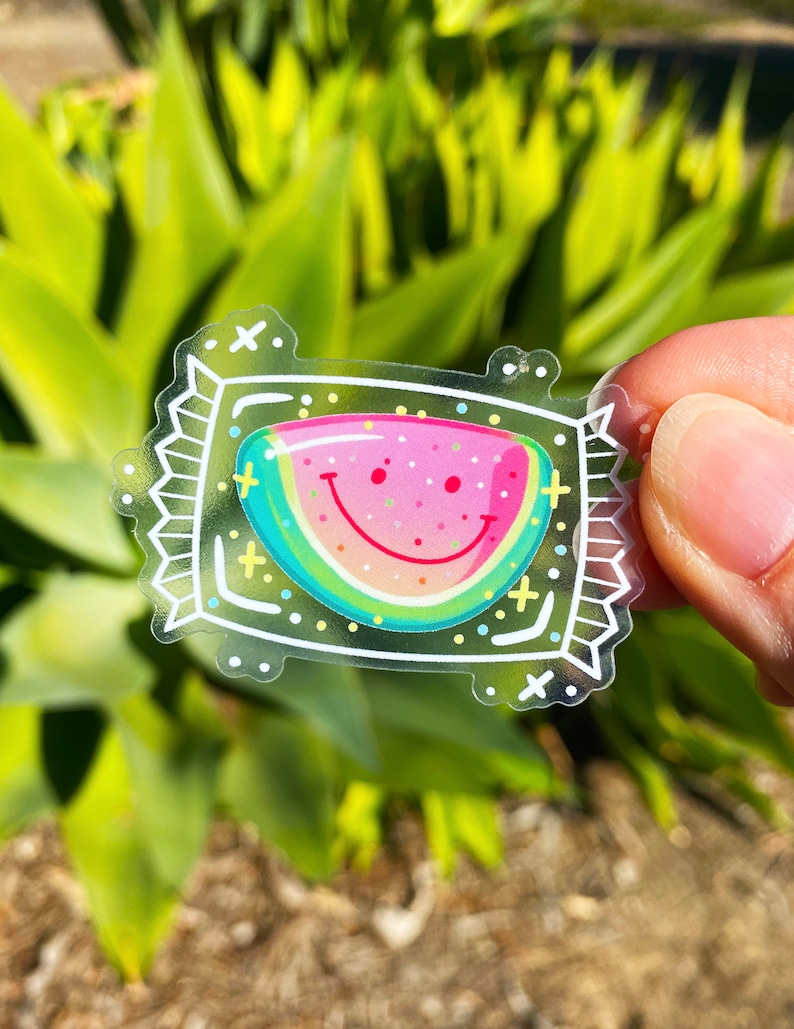 Puede incluir: Una pegatina transparente con un dise&ntilde;o de rodaja de sand&iacute;a. La sand&iacute;a es rosa y verde con una cara sonriente y semillas blancas. La pegatina est&aacute; rodeada de un marco blanco con un contorno negro y elementos decorativos.