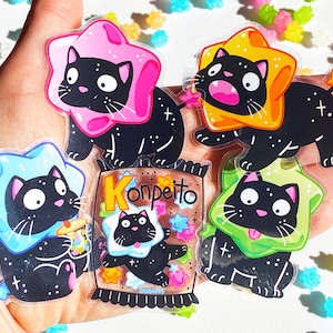 May include: Five black cat stickers with different colored star-shaped hats. The hats are pink, yellow, blue, green, and a brown and white package with the word "Konpeito" on it. The stickers are on a hand.