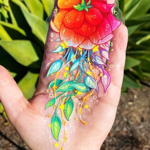 Puede incluir: Una pegatina transparente con una flor roja con hojas amarillas y verdes y estrellas doradas. La flor est&aacute; rodeada por un halo de arco&iacute;ris.