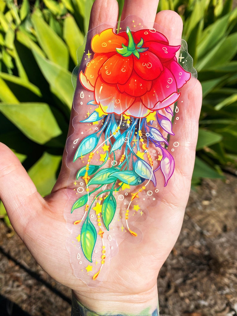 Puede incluir: Una pegatina transparente con una flor roja con hojas amarillas y verdes y estrellas doradas. La flor est&aacute; rodeada por un halo de arco&iacute;ris.