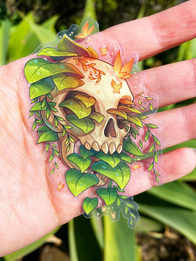 May include: A clear sticker with a white skull surrounded by green leaves and orange butterflies. The skull is partially obscured by the leaves.