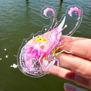 K&ouml;nnte beinhalten: Ein klarer Aufkleber mit einem rosa, garnelenartigen Wesen mit einem l&auml;chelnden Gesicht, einem Stern und einer Erdbeere. Das Design umfasst wei&szlig;e Fl&uuml;gel, Regenbogenakzente und rosa Federdetails. Der Aufkleber wird vor einem Wasserhintergrund gehalten.