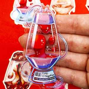 Pu&ograve; includere: Un adesivo trasparente con un design stilizzato di una lampada a lava con liquido rosso e viola. L'adesivo ha un codice a barre e il testo "TYPE: O Negative NON-VOLUNTEER DONOR". Sfondo rosso.