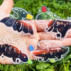 K&ouml;nnte beinhalten: Vier transparente Vinyl-Aufkleber mit Schwarzwei&szlig;-Illustrationen von Dinosauriern mit einem roten, blauen oder gelben Akzent. Die Dinosaurier sind ein Pterodactylus, ein Stegosaurus, ein Triceratops und ein T-Rex.