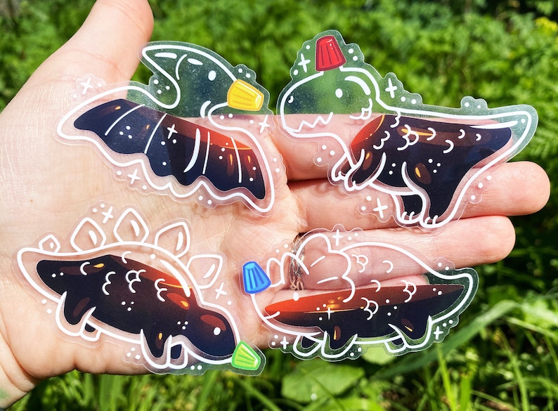 K&ouml;nnte beinhalten: Vier transparente Vinyl-Aufkleber mit Schwarzwei&szlig;-Illustrationen von Dinosauriern mit einem roten, blauen oder gelben Akzent. Die Dinosaurier sind ein Pterodactylus, ein Stegosaurus, ein Triceratops und ein T-Rex.