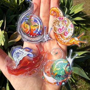 May include: A hand holding five clear stickers of whimsical snails. Each snail features a unique shell design with vibrant colors, including floral patterns, gems, and intricate details. The stickers are transparent, allowing the background to show through.