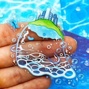 May include: A clear sticker featuring a whimsical island scene. The island has a green grassy top with buildings and a blue lake. A blue butterfly and bubbles surround the island. The sticker is held in a hand.