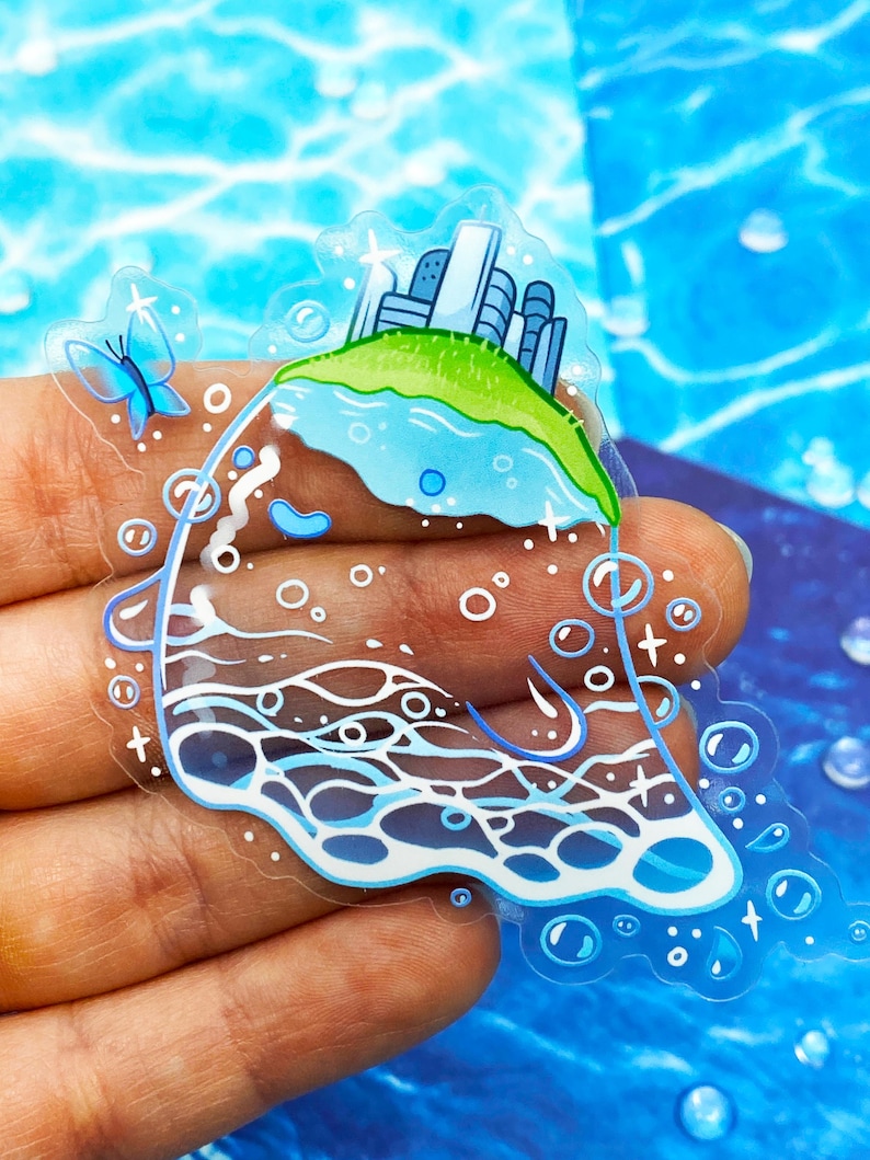 May include: A clear sticker featuring a whimsical island scene. The island has a green grassy top with buildings and a blue lake. A blue butterfly and bubbles surround the island. The sticker is held in a hand.