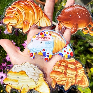 May include: Four colorful stickers featuring cartoon manatees with different food-themed designs. The manatees are depicted as a croissant, a box of Wonder bread, a stack of pancakes, and a bowl of ice cream.