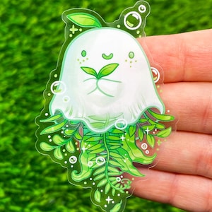 May include: A clear sticker featuring a whimsical white ghost-like character with green fern-like details. The character has a smiling face with green leaves and is surrounded by bubbles and leaves. The sticker is held against a green background.